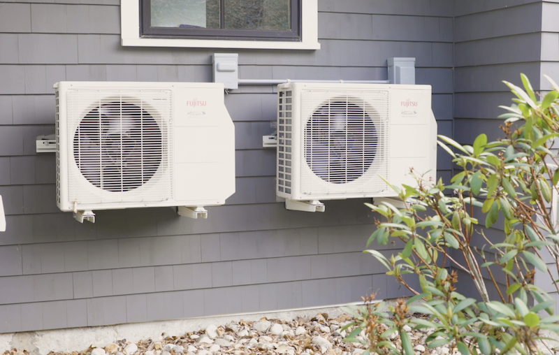 Heat pumps on the outside of a grey, wooden home while it is sunny outside.