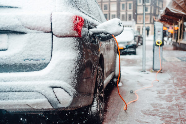 A view of half an electric car, from its trunk, covered in snow charging on the sidewalk.