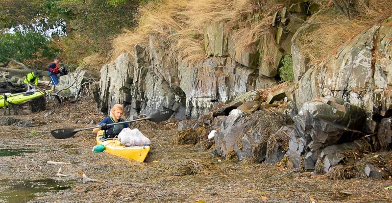 Instructor from Seven Rivers Paddling rides a yellow kayak by the shoreline of the Great Bay Estuary. On the kayak, her carries a mesh bag with trash from the cleanup.