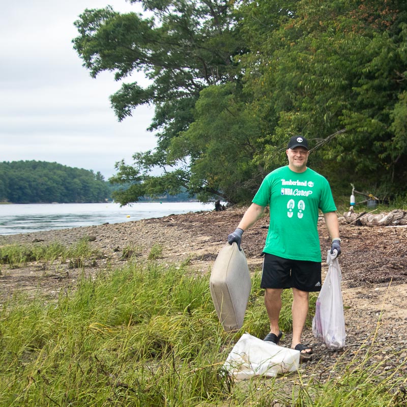 Cleanup volunteers collecting trash on the shoreline of the Great Bay estuary. On left hand, he holds a cushion. On his right hand, he hold a mesh bag with marine debris.