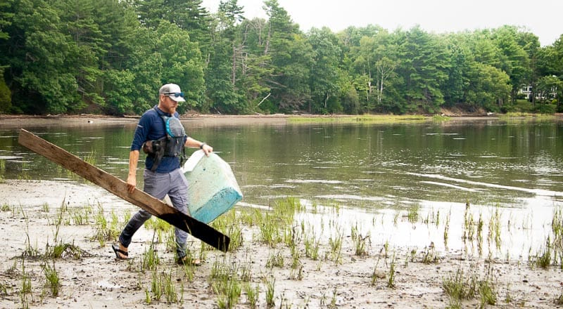 Peter Sawtell from Seven Rivers Paddling holds a large block of blue dock foam and a piece of rotten wood he found during the cleanup. On the background, the Great Bay estuary gleams with the midday sun.