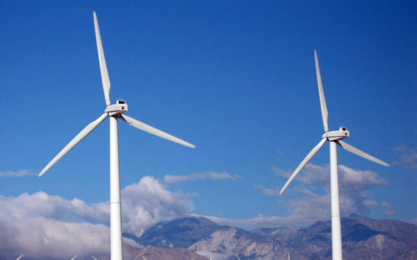 Two wind turbines, which harness wind power as a source of renewable energy, against a cloudy sky with mountains in the distance.