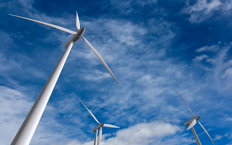 A view from the ground-up of wind turbines against a blue sky with some clouds.