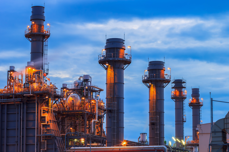 A photo of a gas power plant against a deep blue sky. The infrastructure is lit by an orange glow.