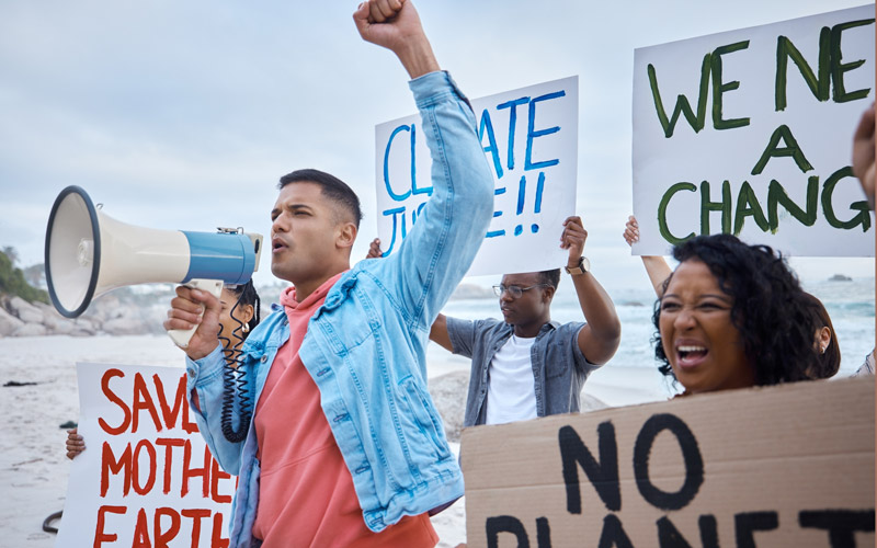 Group of multiracial activists call out for climate justice while holding signs and a megaphone. In the background, a sandy beach and the ocean.
