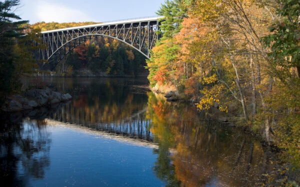 An angled view of the French Kiung Bridge, Erving, Massachusetts. The bridge reflects into the body of water below, surrounded by trees that are changing colors for the fall.