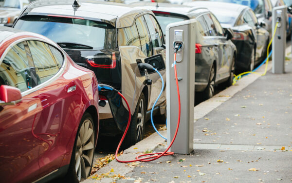 A row of cars alongside a curb plugged into curbside charging stations to recharge while parked. A red sedan is first in view, and then a black/grey SUV behind it.