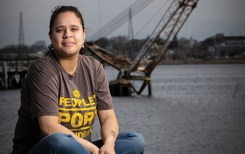 Monica Huertas, a Latina woman wearing a brown t-shirt and jeans, sitting in front of the water with a crane in the background. Her shirt reads "People's Port Authority."