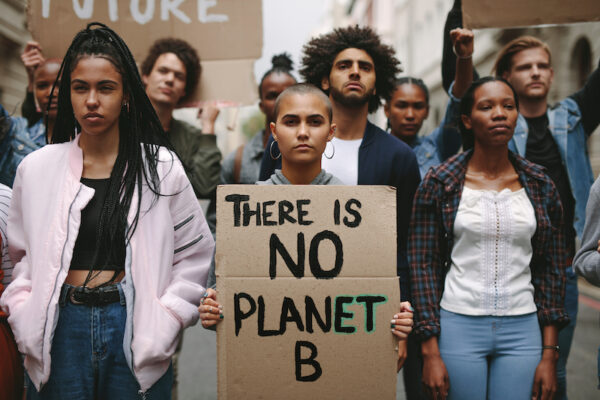 A group of people facing the camera. A young person stands in the center directly looking into camera holding a sign that says "There is no planet b." They are surrounded by other young adults, some of which are holding other signs (slightly out of focus).
