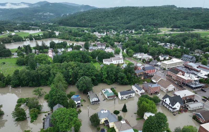 This aerial photo shows flooding in a neighborhood in Montpelier, Vermont. Brown water covers the streets and yards of homes and businesses. Green mountains rise in the distance. Photo credit: Vince Franke