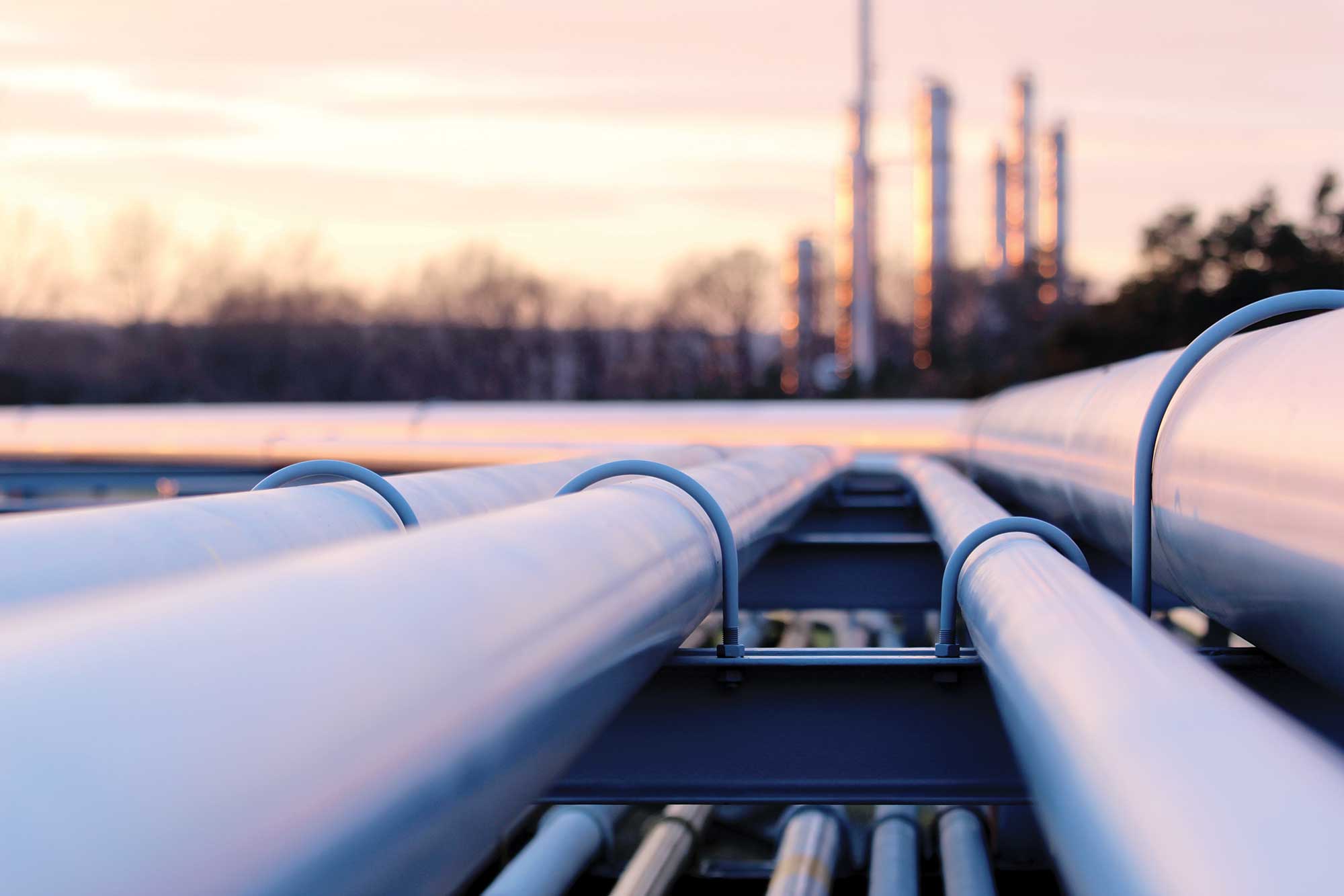 Photo shows four pipes from a natural gas facility with the towers of the gas plant blurred in the background, against a sky colored pink and orange from the sunset. Credit: Shutterstock