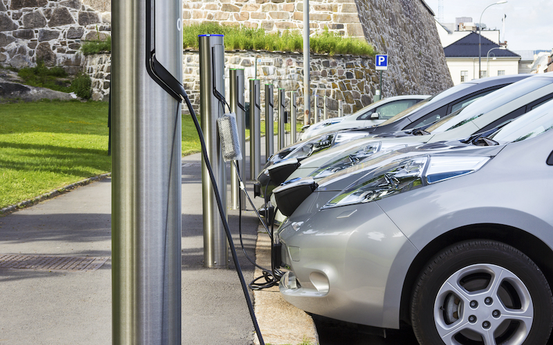 a row of grey/silver electric cars parked against a curb charging. You can only see the front of the car up to half the front tire.