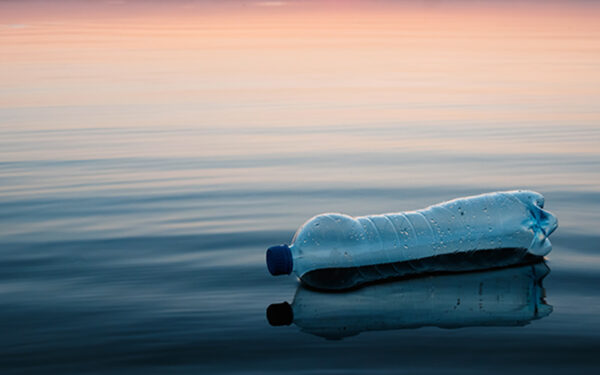 Empty single-use plastic bottle sits on shore over water. Photo illustrates that plastic products do not break down or biodegrade when in contact with natural elements. From top to bottom, the background shows a gradient going from light orange to white to dark blue. These sunset-like colors are reflected in the water.