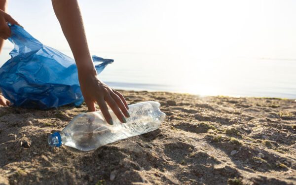 Close-up of an empty plastic bottle being picked off from the ground by person wearing blue nail polish. In the background, there's sand, the ocean, and a bright sunny sky.