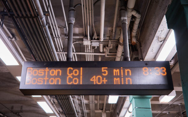 An electronic board shows arrival times for train cars, one approaching in 5 min and another one approaching in 40+ minutes as a result of MBTA slow zones. In the background, you can see a green column, indicating that this photo was taken at Green Line station. Metal pipes and white fluorescent lights on the ceiling.