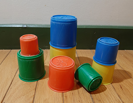 Plastic stacking cups in colorful tones of blue, green, orange, and yellow. Colors are muted and cups look scratched, used, and old. Imprinted at the bottom of the cups, whimsical figures of animals are imprinted. Cups sit on a light wooden floor in front of a white wall with a dark green baseboard at the bottom.