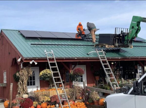 A view of two people installing solar panels onto the green roof of a burgundy building, adorned with hanging flowers, pumpkins in wooden baskets, and white doors.