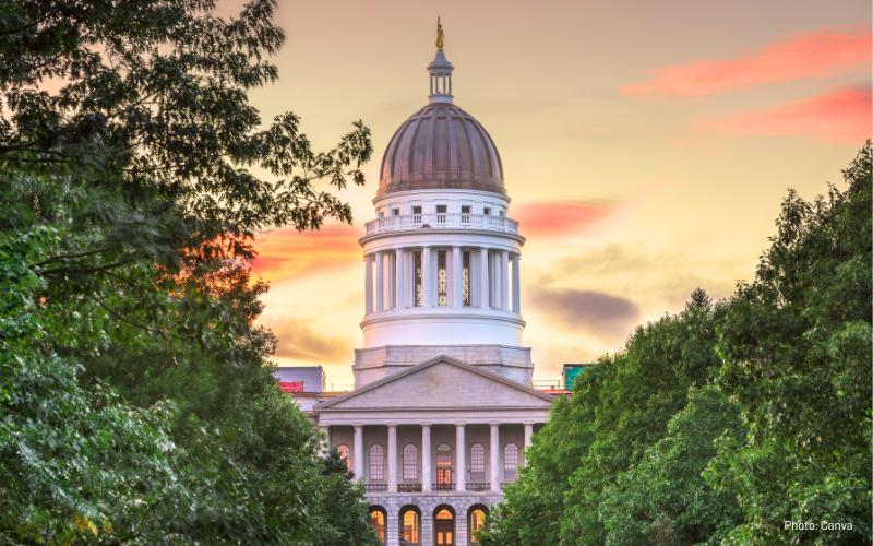 Top of Maine State House over sunset, green lush trees on the left and right of the foreground.