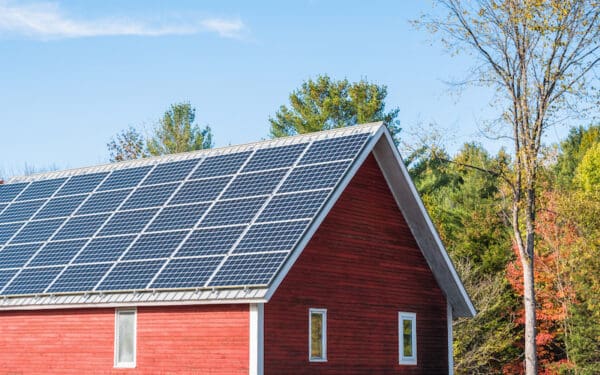 A view of the top half of a red barn with solar panels ovr the entire roof. The barn is against a blue sky with trees changing to their fall foliage color.