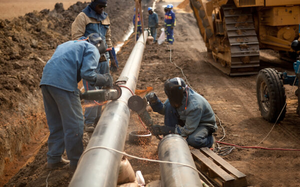 Two construction workers in blue jumpsuits wearing masks are bent over welding a large pipeline. The pipeline stretches far into the background against a dug-out dirt background, where more construction workers are seen in the distance.