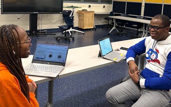 Two people talk at a table with laptops