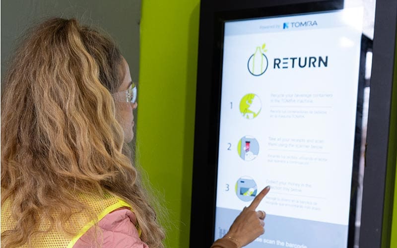 Alaina Pinto standing at a bottle return machine and pointing at the instructions on the touchscreen inside a recycling center.