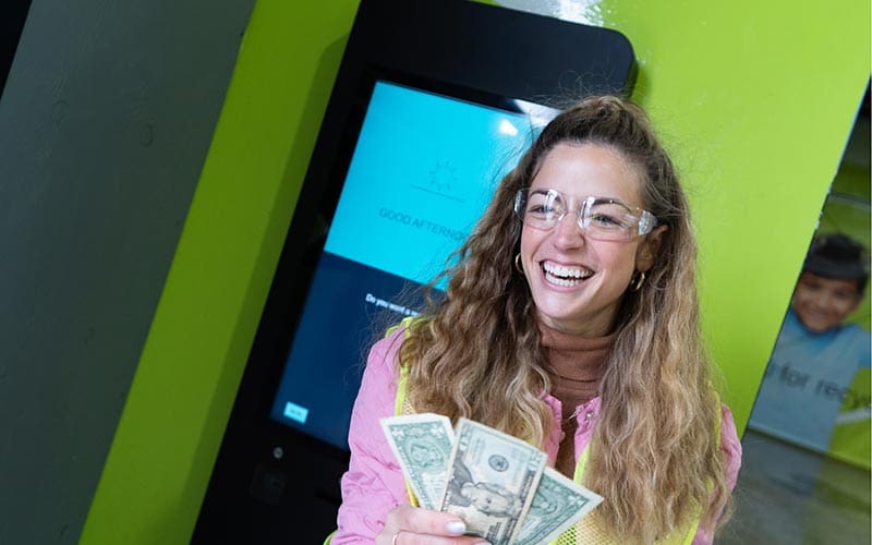 Alaina Pinto smiling and holding cash in her hand while standing in front of a bottle return machine at a recycling center.