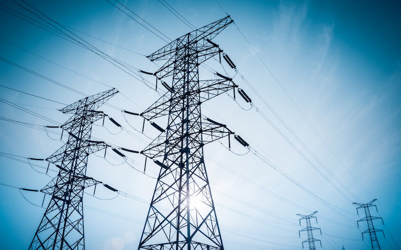 At an angle facing upwards, against a blue sky, are two electricity transmission towers.