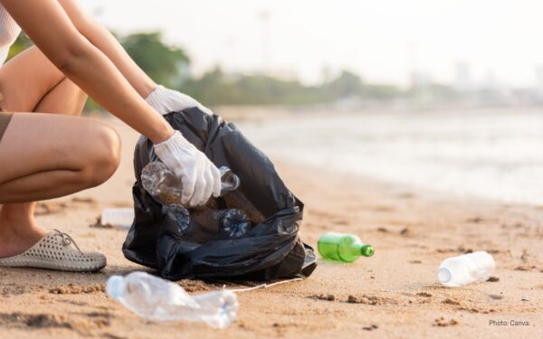 Sunny beach day. A person wearing white gloves picks up empty plastic bottles from the sand and puts them inside a black plastic bag as part of a community cleanup. Cleanups like this help document the impact of plastic litter on our environment.
