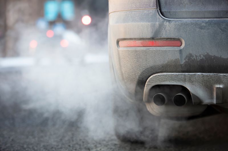 a close up of a silver car's tailpipe. the car is driving on a road with other cars, blurred in the background. The tailpipe has grey fumes and smoke coming out of it, concealing most of the background.