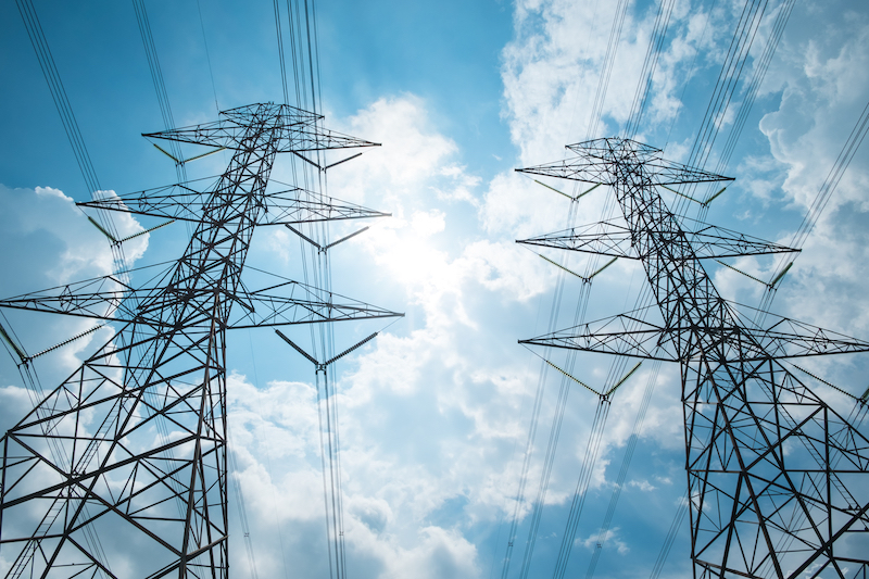 A bottom-up view of transmission poles and wires against a cloudy, but sunny, sky