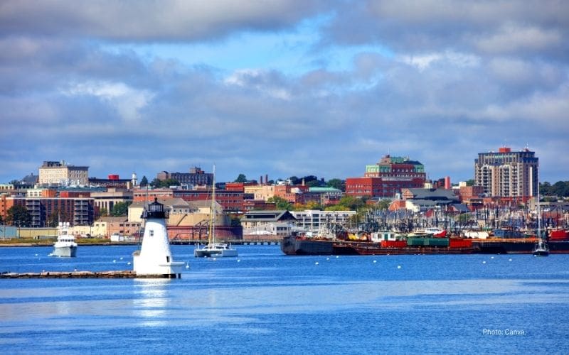 View of New Bedford harbor with a white lighthouse in the foreground, sailboats on the water, and the city skyline with colorful buildings under a partly cloudy sky.