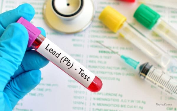 Close-up of a gloved hand holding a blood vial labeled "Lead (Pb) - Test," surrounded by test tubes, a syringe, and a stethoscope on a medical report – illustrating how lead poisoning is diagnosed through blood testing.