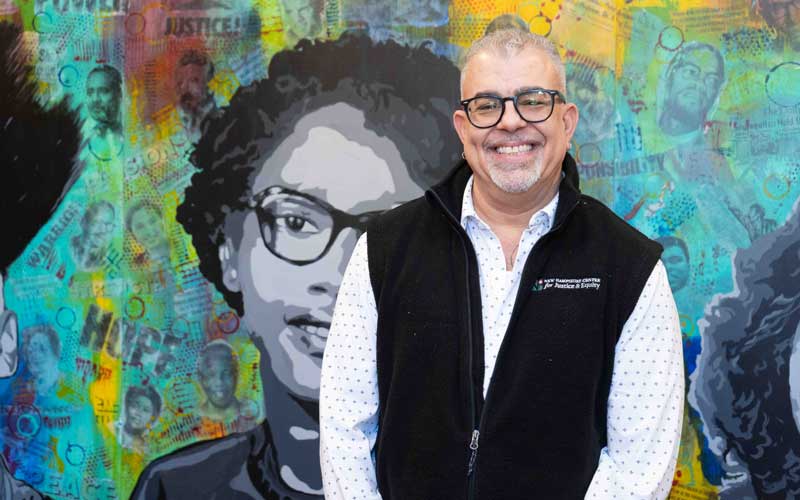Anthony Poore stands smiling in front of a colorful mural featuring civil rights imagery. He wears a vest with the New Hampshire Center for Justice & Equity logo.