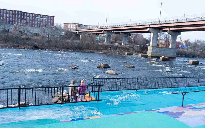 View of the Merrimack River on a cold day, with two people standing on a painted riverside walkway and a bridge in the background.
