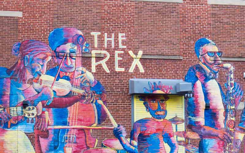 A vibrant mural painted on the red-brick exterior of The Rex Theatre shows musicians playing instruments, expressing energy, culture, and community.