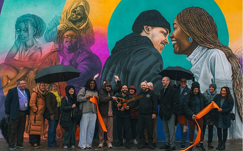 A diverse group of people pose in front of a colorful mural during a ribbon-cutting ceremony on a rainy day, with some holding umbrellas and orange ribbon.