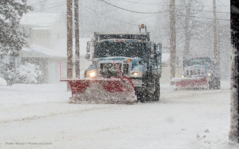 Two blue trucks, one in the foreground and one out of focus in the background, plow snowy roads with red plows. It is snowing heavily in a residential neighborhood.