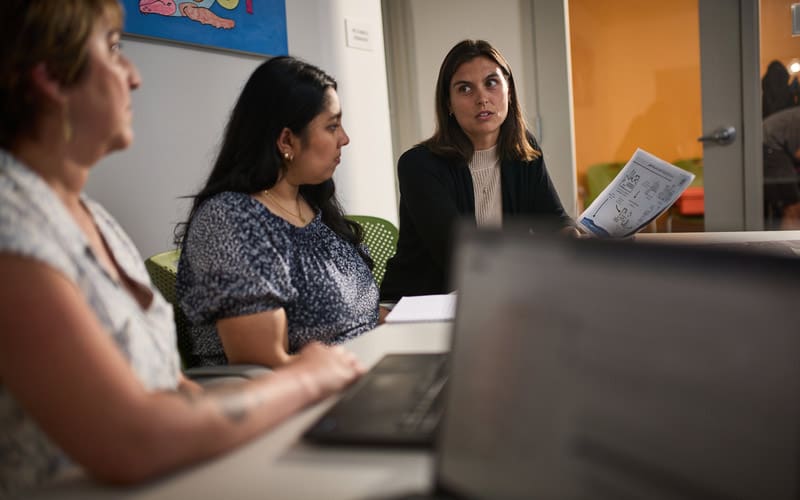 Three women sit at a conference table during a meeting, one holding a printed document. A laptop is in the foreground, and colorful artwork hangs on the wall behind.