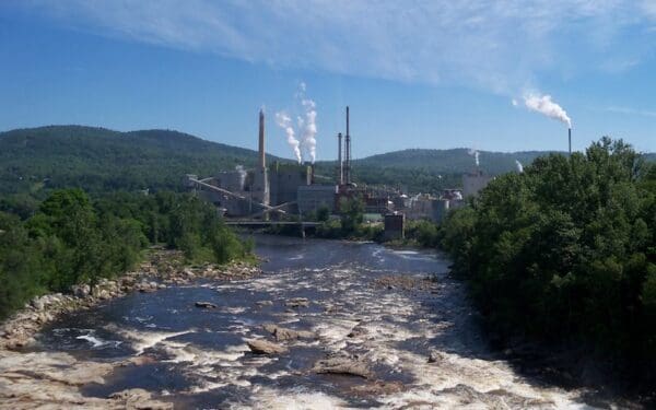A view of Rumford Paper Mill in the distance across a streaming river. There is smoke coming out of the Mill.