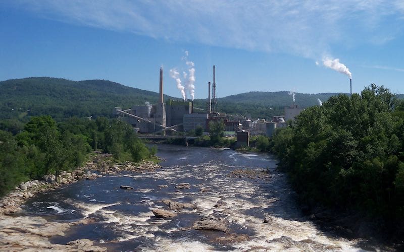 A view of Rumford Paper Mill in the distance across a streaming river. There is smoke coming out of the Mill.