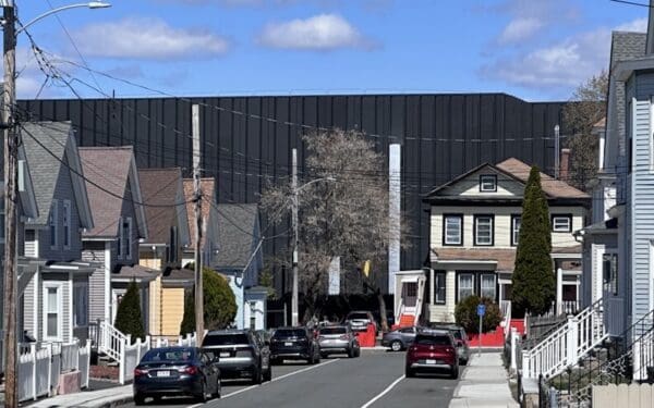 Photo of two-story neighborhood houses along a street in Lowell, Massachusetts, with the black facade of a data center looming over the homes in the background.