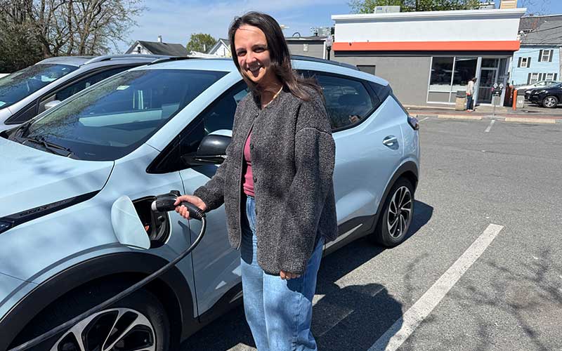 Emma Keaney stands next to her sky blue electric vehicle while holding a charger plugged into her car.